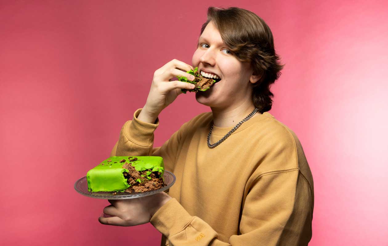 Ruskeaan collegepaitaan pukeutunut Kakkuh pitää kädessään tarjoilulautasta, jolla on vihreä suklaakakku. Kakkuh on juuri repinyt käsillä palan irti ja pitelee sitä suussaan samalla kuin hymyilee kameralle. 