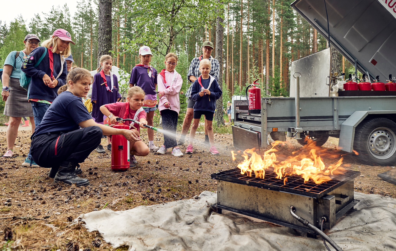 Partioleirillä leiriläiset pääsevät sammuttamaan tulipaloa. Yksi leiriläisistä sammuttaa tulipaloa ja aikuinen opastaa häntä vieressä. Muut seuraavat kuvassa takana.