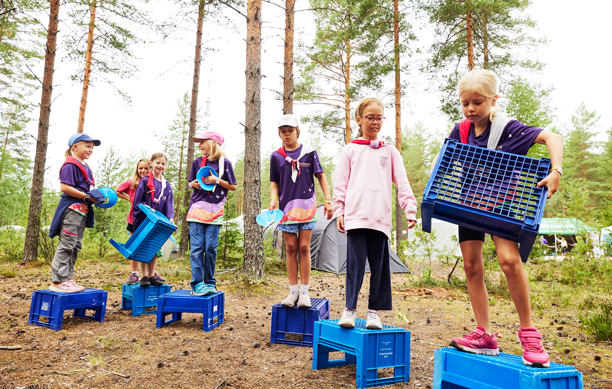 Kuvassa kuusi partiolaista seisoo maitokorien päällä metsässä.