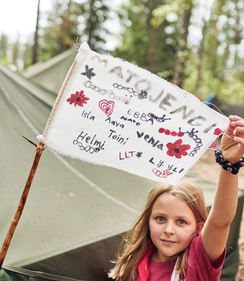 Luotsitytöistä yksi esittelee partioleirillä lippua, jossa lukee matojengi ja sen jäsenten nimet. 